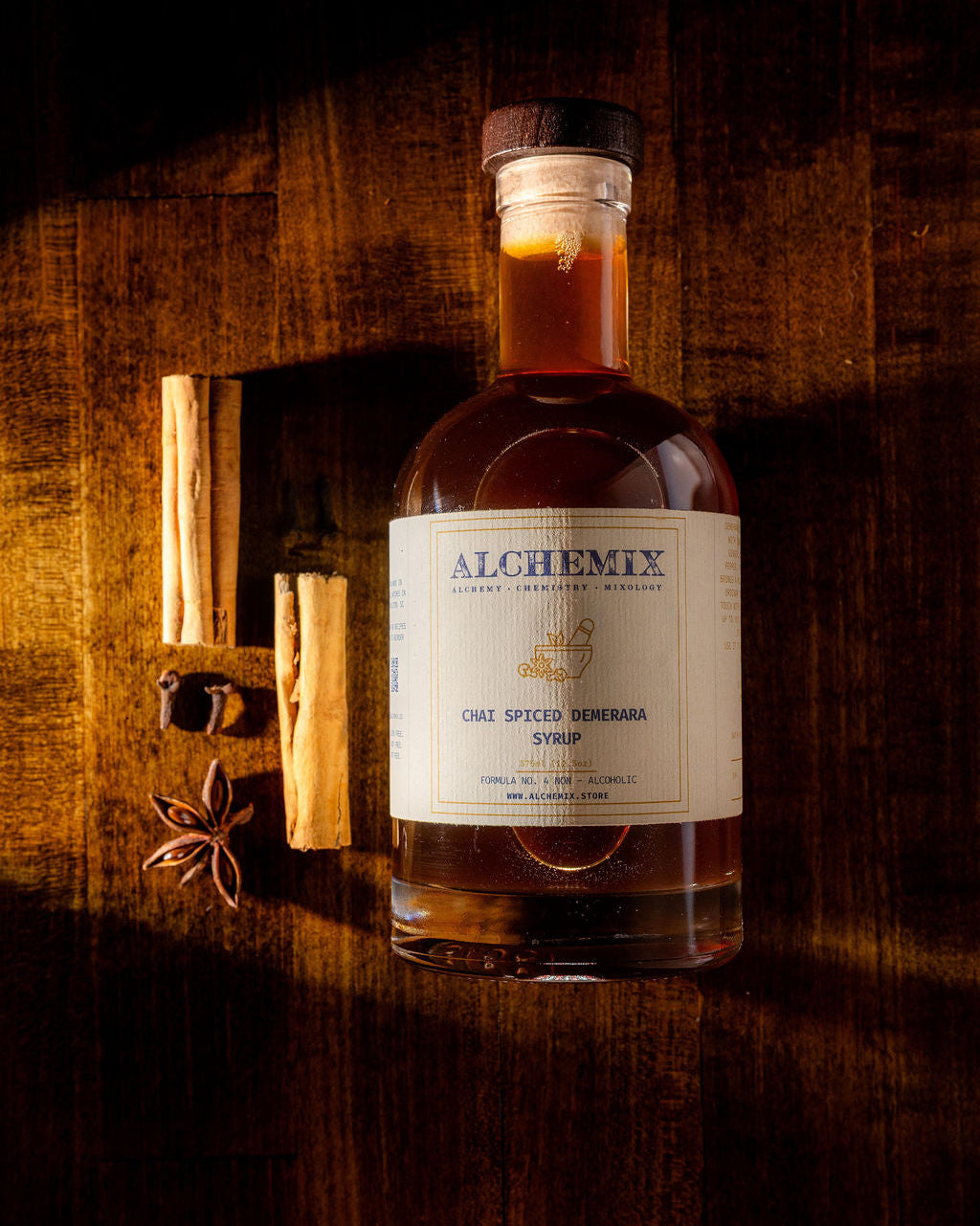 A bottle of Alchemix Chai Spice Demerara Syrup lays on a dark wooden surface with warm, dramatic lighting. The bottle is filled with amber liquid and has a cream-colored label with blue text.