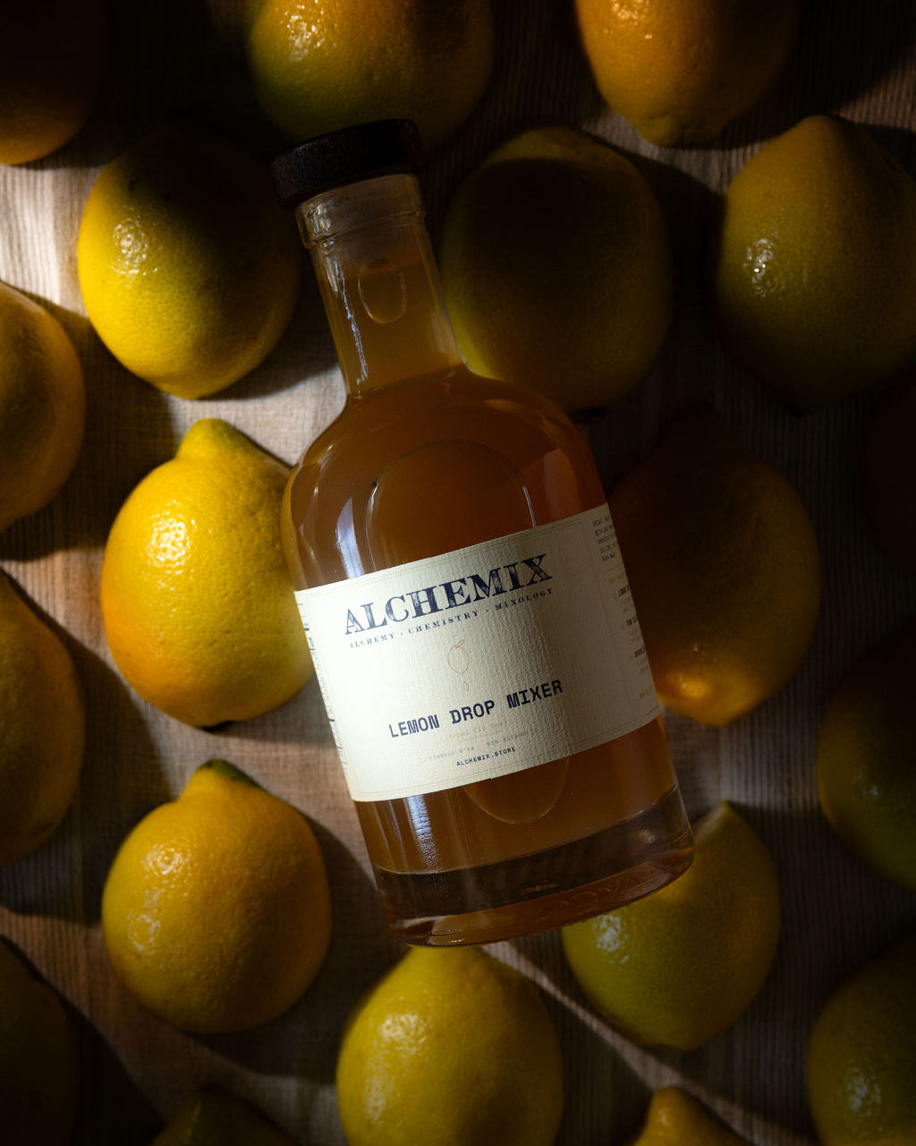 A bottle of Alchemix Lemon Drop Mixer surrounded by fresh yellow lemons on a dark wooden surface.