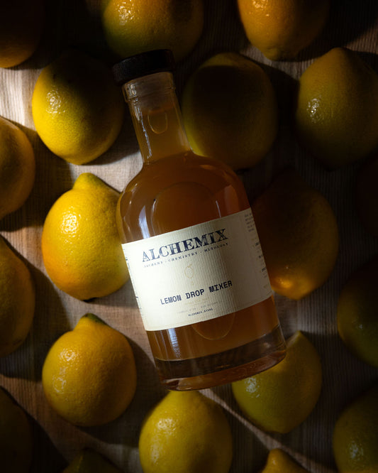 A bottle of Alchemix Lemon Drop Mixer surrounded by fresh yellow lemons on a dark wooden surface.