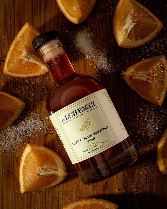 A bottle of Alchemix Lightly Salted Grapefruit Syrup lies on a wooden surface, surrounded by grapefruit slices and scattered coarse salt. The bottle has a cream label with  text and a black cap.