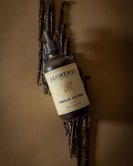 A bottle of Alchemix Vanilla Bitters with a black dropper cap and cream label, styled diagonally among dried vanilla pods on a warm brown background.