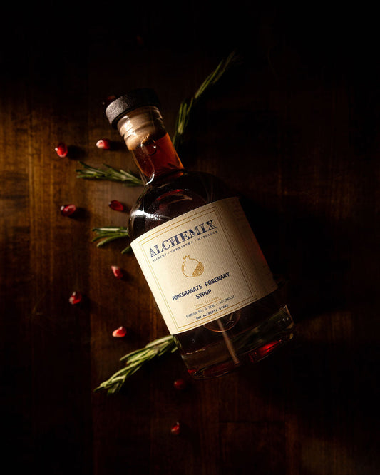 A bottle of Alchemix Pomegranate Rosemary Syrup lies on a dark wooden surface, surrounded by rosemary sprigs and scattered red berries, in dramatic low light.